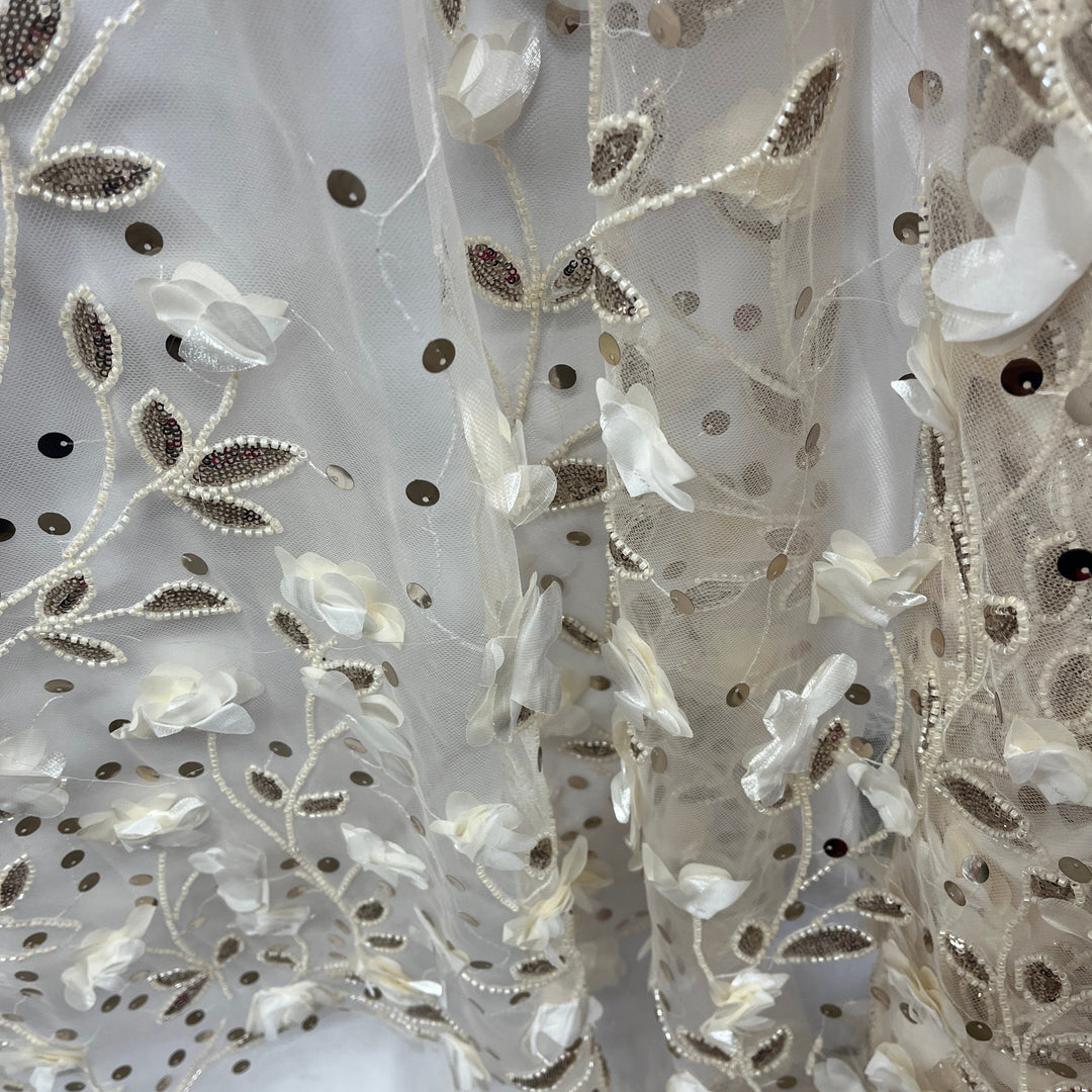 Close-up of a sheer white tulle fabric with embroidered floral and leaf patterns in white, pearl-bead, and metallic Champagne tones.