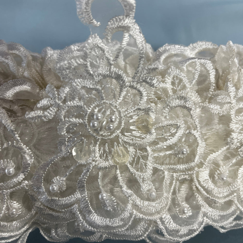 Close-up of an intricate ivory lace fabric with floral embroidery, bead details,  against a soft blue-gray background. LaceUSA #KZ-50