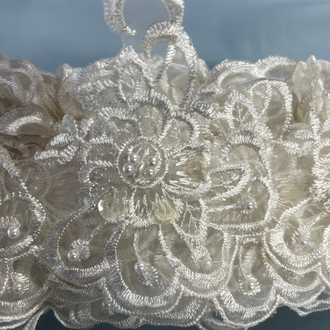 Close-up of an intricate ivory lace fabric with floral embroidery, bead details,  against a soft blue-gray background. LaceUSA #KZ-50