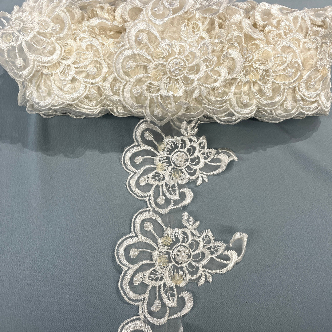 Ivory floral lace trim embellished with embroidered flowers, pearls, and sequins, featuring a scalloped edge and intricate floral pattern., draped over a gray fabric background. LaceUSA #KZ-50