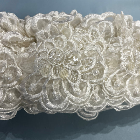 Close-up of an intricate ivory lace fabric with floral embroidery, bead details,  against a soft blue-gray background. LaceUSA #KZ-50