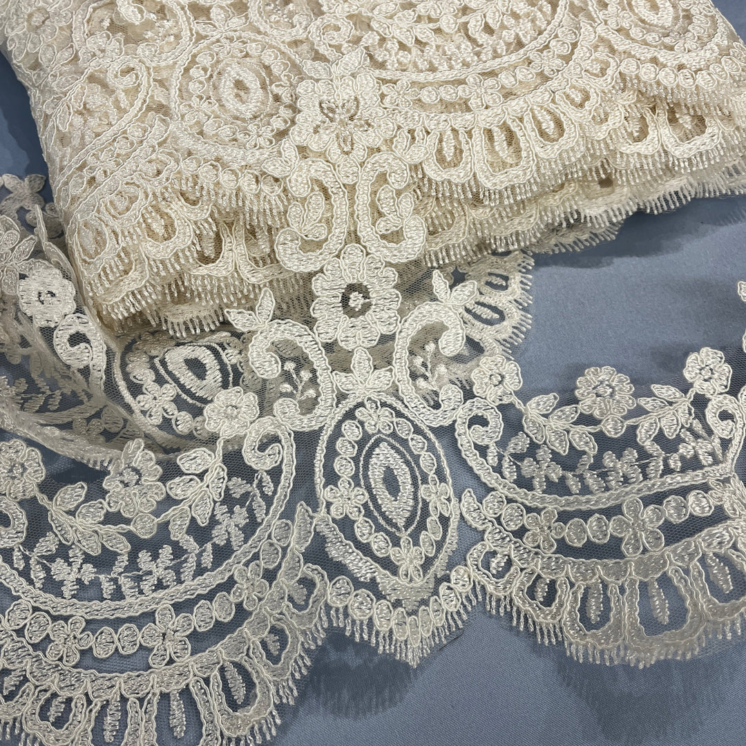 Close-up of ivory floral lace trim with ornate embroidery and scalloped edging, laid on matching lace against a slate blue background, Lace USA 96794W.