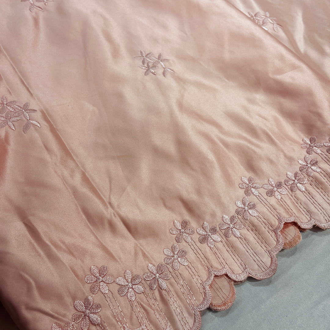 A close-up of a peach satin fabric with delicate floral embroidery and a scalloped embroidered border, laid out on a pale grey surface.