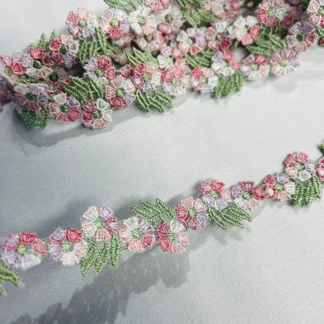 A close-up of a floral embroidered lace trim featuring  a multicolor pink, white, and light purple flowers with green leaves, laid out on a light gray fabric background.