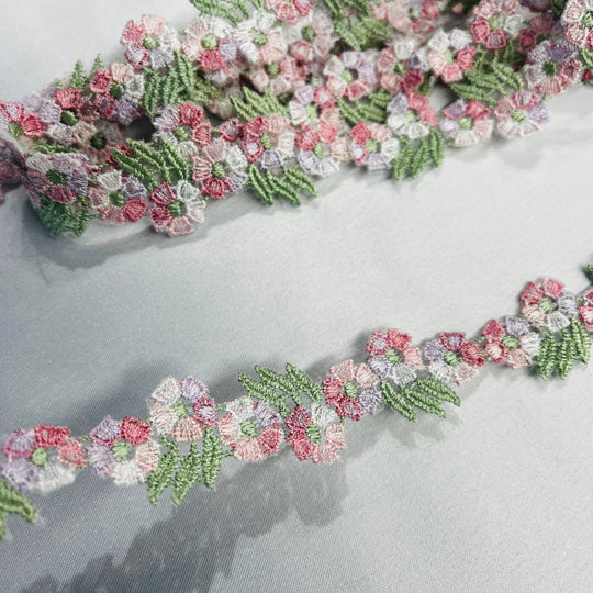 A close-up of a floral embroidered lace trim featuring  a multicolor pink, white, and light purple flowers with green leaves, laid out on a light gray fabric background.