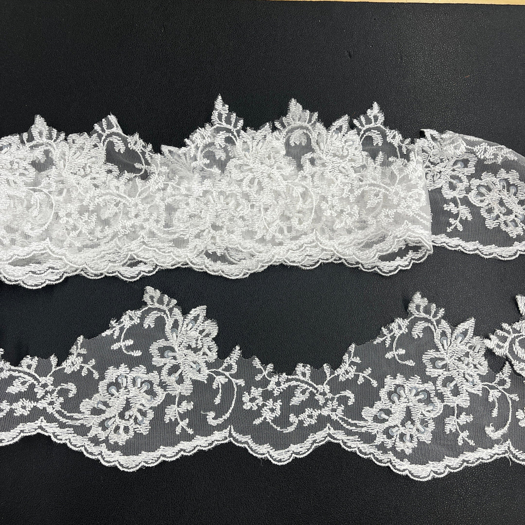 White beaded floral lace trim with scalloped edges, displayed in two horizontal strips featuring one flat unrolled length and one gathered bundle on a black background. Lace_USA #21356WBP