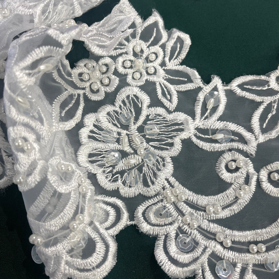 Closeup of White floral lace fabric with embroidered on polyester organza  embellished with pearl beads and sequins,  shown in a close-up against a dark green background. Lace USA  #890408NBP
