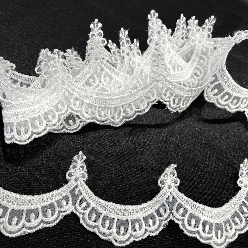 Closeup of White lace trim with a decorative floral and scalloped edge, shown in two curved pieces on a black background. Lace USA 21618N
