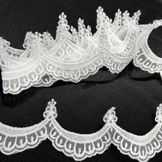 Closeup of White lace trim with a decorative floral and scalloped edge, shown in two curved pieces on a black background. Lace USA 21618N