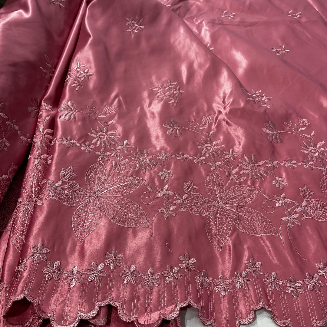 A close-up of a dusty rose satin fabric with intricate floral and leaf embroidery and a scalloped border.