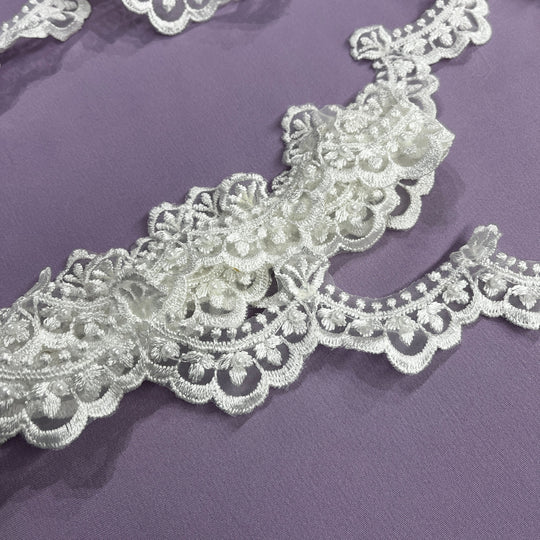 Closeup of Off White Ivory embroidered lace trim on organza embellished with pearls and sequins with floral and scalloped patterns laid on a solid purple background. LaceUSA  Item 20648NBP 