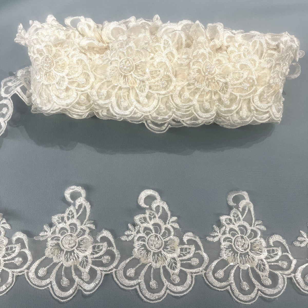 Ivory floral lace trim with intricate embroidered flowers, pearl and sequin embellishments, scalloped edges, and a matching folded lace piece above on a dark gray background. LaceUSA #KZ-50