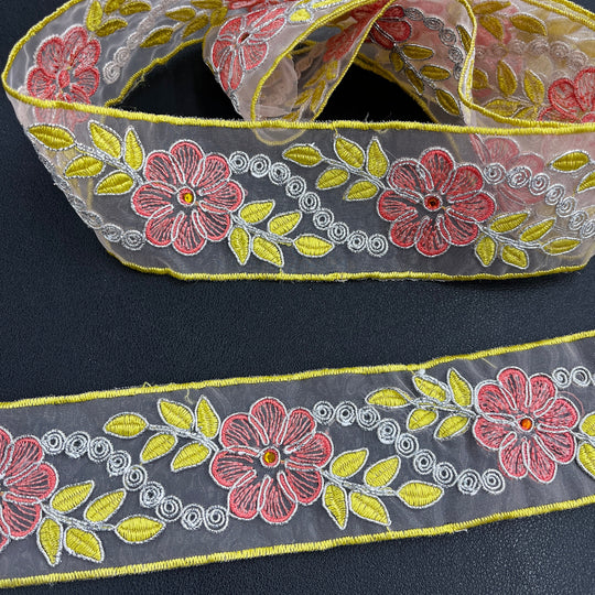 Close-up of two pieces of the same decorative embroidered lace trim, featuring pink flowers with rhinestone centers, yellow leaves, and silver scroll details, corded with metallic silver thread on sheer organza fabric over a dark surface. Lace USA #96307NRS. 
