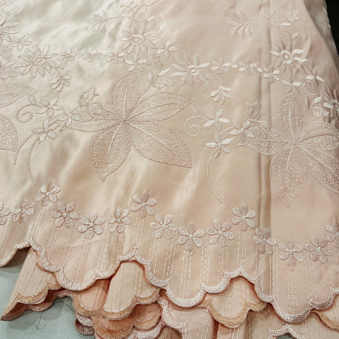 A close-up of a peach satin fabric with intricate floral embroidery and a scalloped embroidered border.