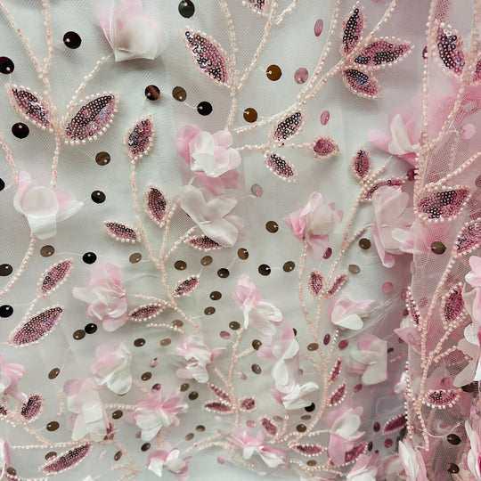 A close-up of a light pink and white floral embroidered fabric with 3D flower appliqués, beaded vines, and sequined leaves.