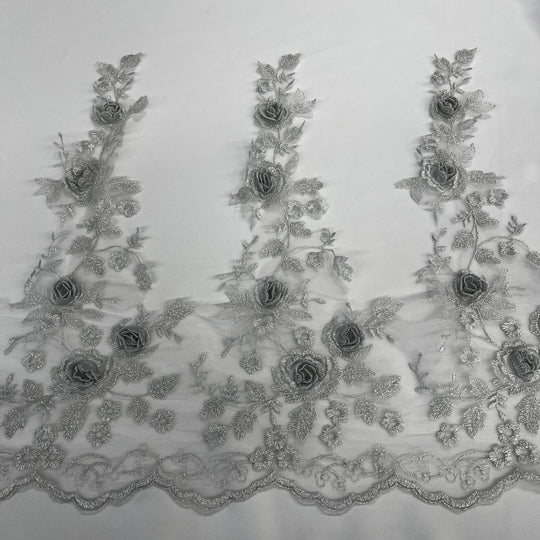 Silver floral embroidered lace trim featuring 3D rose appliqués, metallic thread embroidery, and a scalloped edge embroidered on net mesh., shown laid flat against a white background. LaceUSA #21854W
