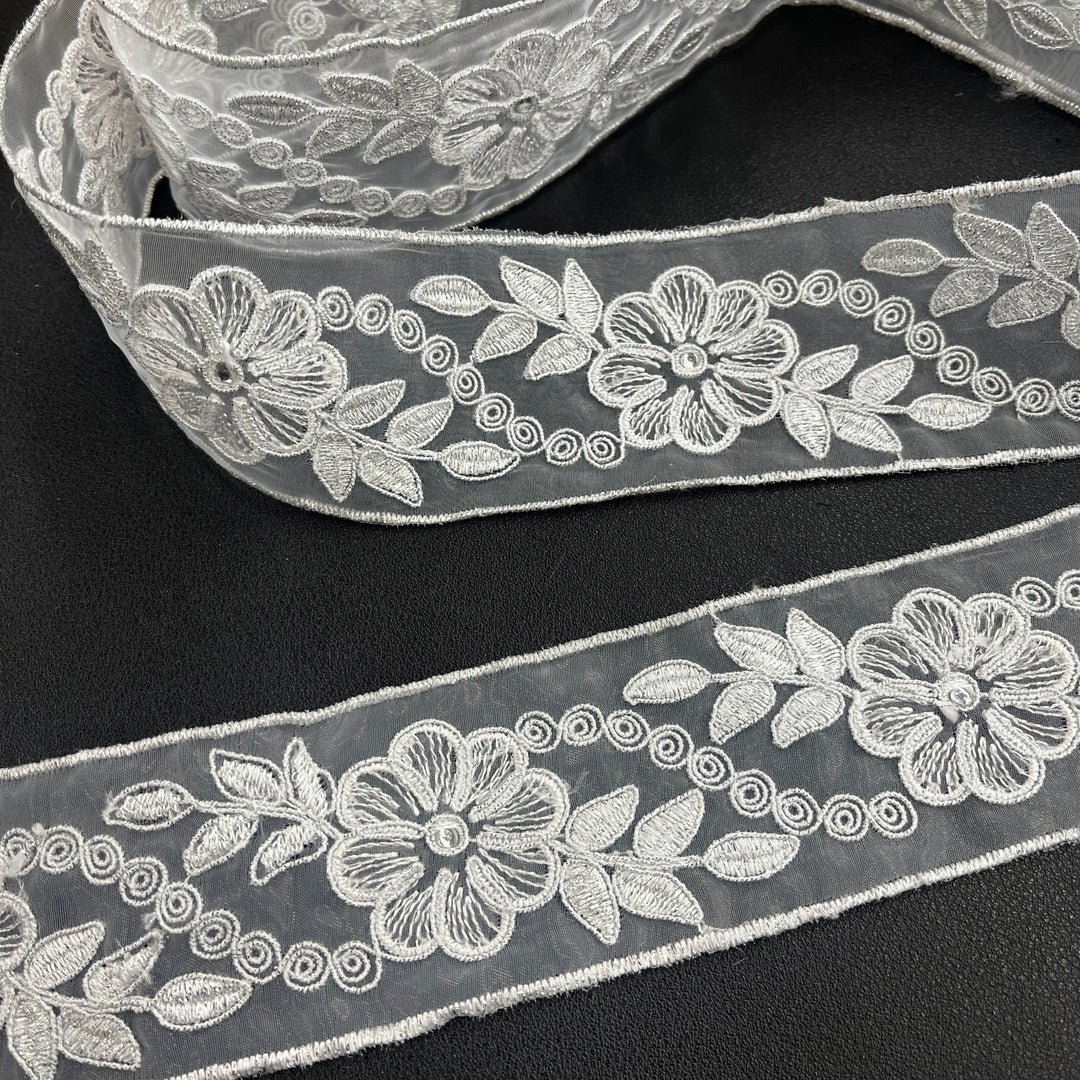 Close-up of two pieces of the same  silver decorative embroidered lace trim, featuring flowers with rhinestone centers, leaves, and scroll details, corded with metallic silver thread on sheer organza fabric over a dark surface. Lace USA #96307NRS. 