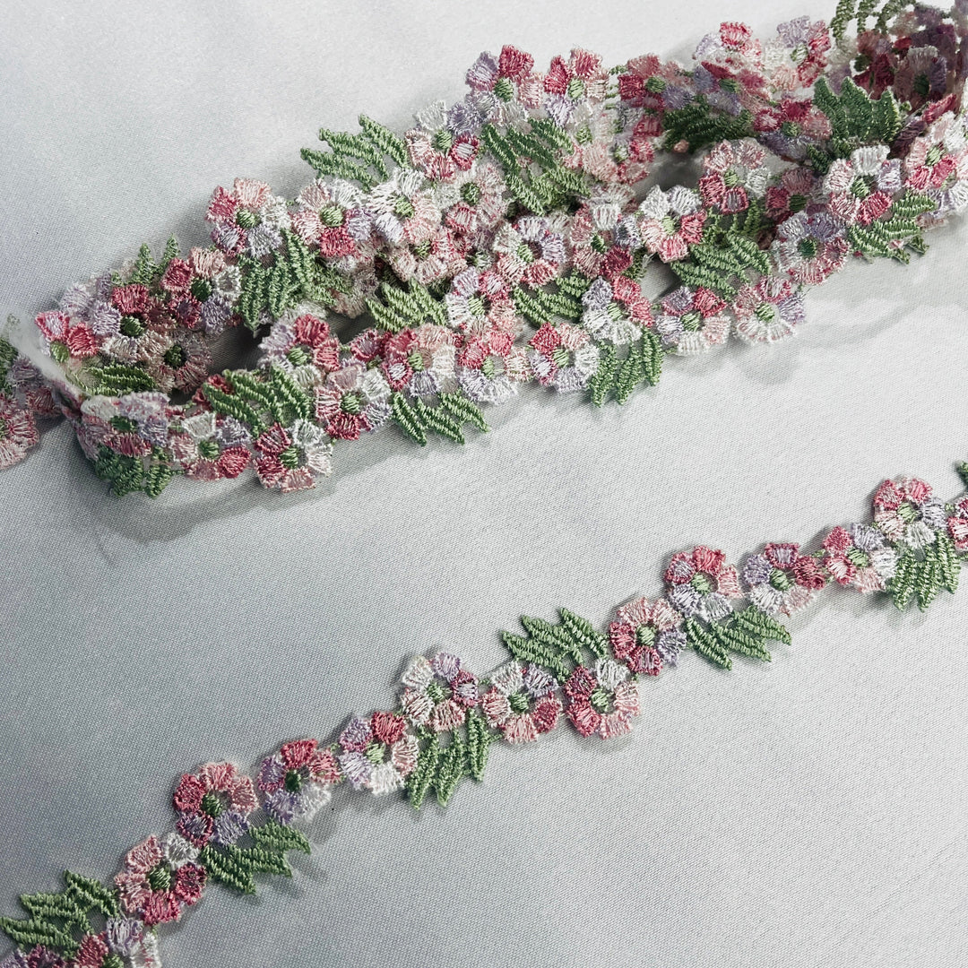 a decorative embroidered lace trim featuring pink and lilac flowers with sage green leaves, laid diagonally on a light gray fabric background. LaceUSA item 20441N