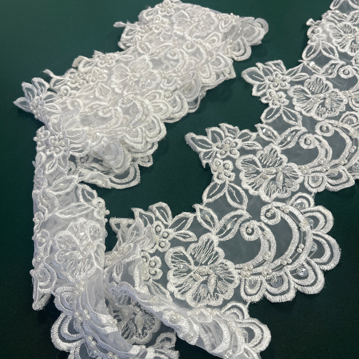 White Beaded floral lace trim with embroidered details and scalloped edges, laid out on a dark green background. Lace USA #890408NBP