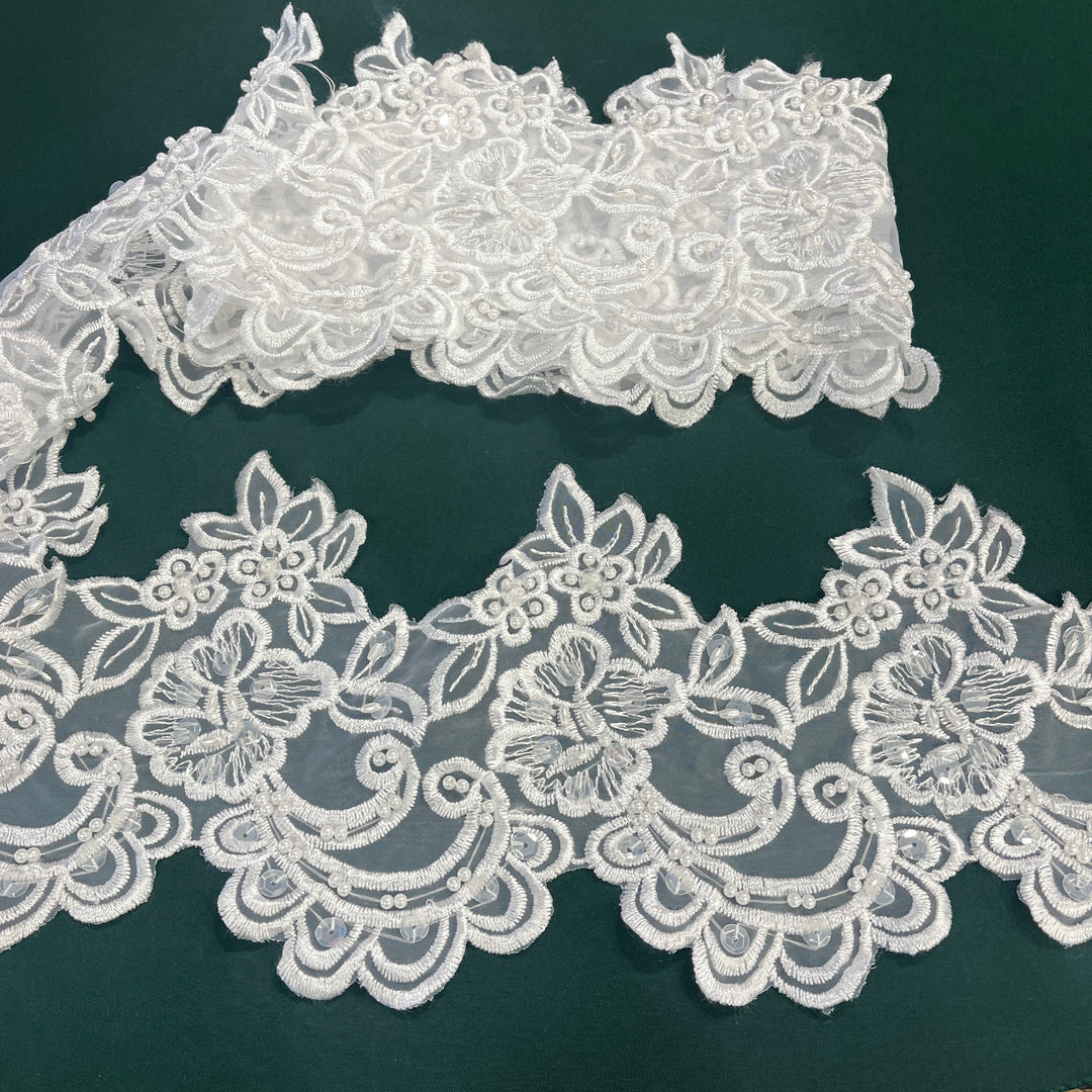 White floral lace trim with intricate embroidered patterns and scalloped edges, embelished with beads & sequins, displayed on a dark green background. Lace USA  #890408NBP