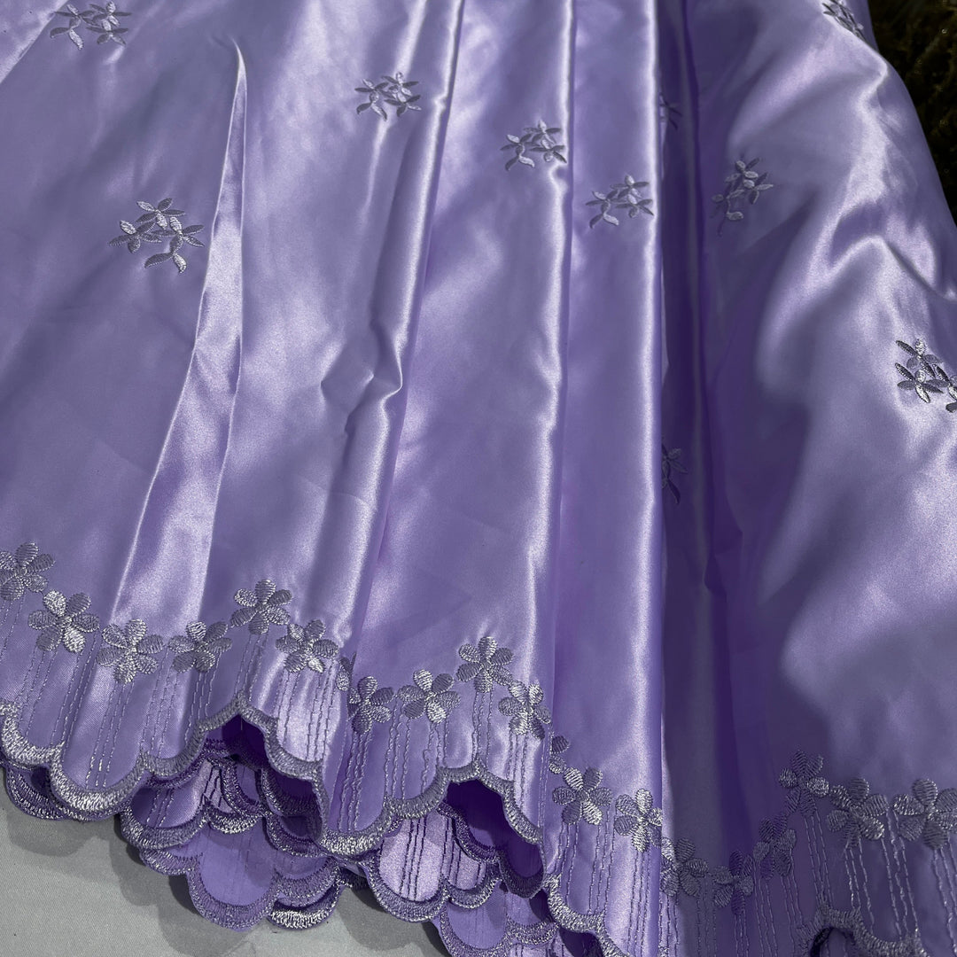 A close-up of a lilac satin fabric with embroidered  floral motifs and a scalloped embroidered hem.